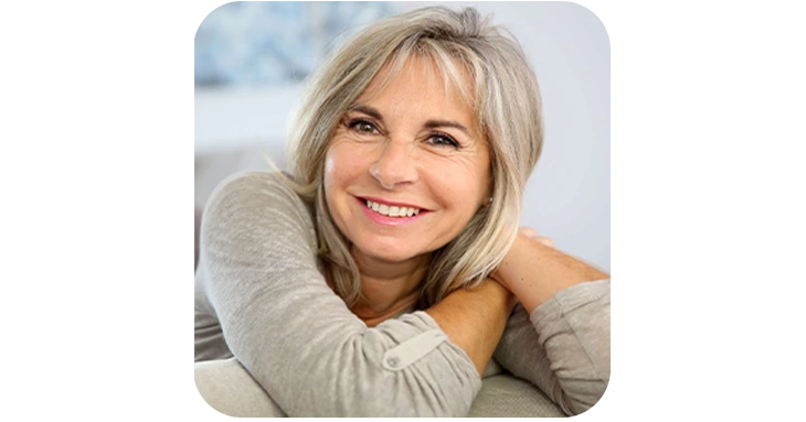 Tonic Greens customer testimonial portrait featuring a smiling, mature woman with blonde-streaked hair resting her arms comfortably on a couch, reflecting vibrant health and vitality.